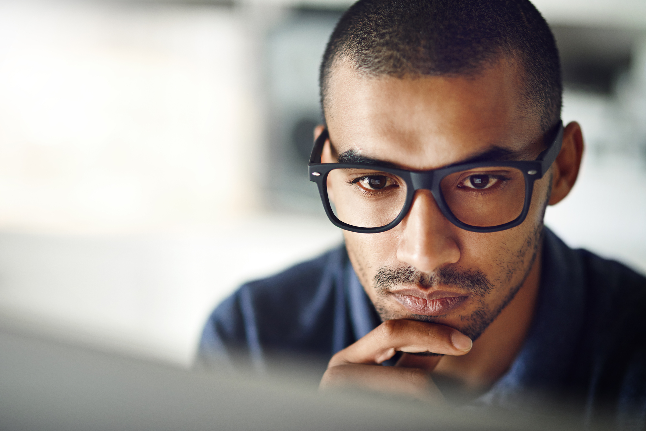 young man wearing glasses looking at a computer screen while deep in thought